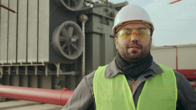 A portrait of a middle-aged power engineer, with a slight unshaven, with overalls, in a protective helmet and glasses against the background of a powerful high-voltage transformer on the territory of