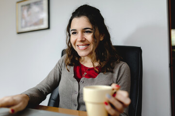 Close-up attractive smiling woman using computer working at office desk with a cup of hot drink.