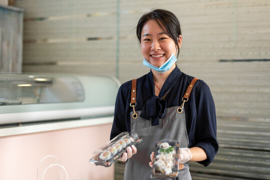 Asian Waitress Shows Traditional Japanese Food Ready In A Plastic Takeout Box, Young Chinese Woman In Uniform At Restaurant Prepares Takeout Bag, Crisis Concept For Coronavirus And New Business