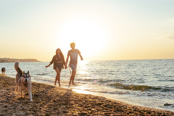 young caucasian couple walking on beach with siberian husky dogs during beautiful sunset or sunrise. Playing with dogs. Pets are the members of family concept