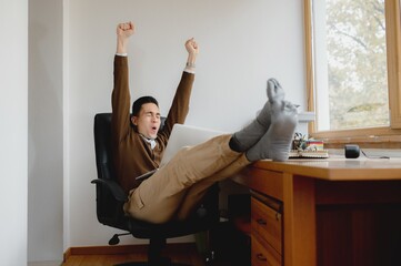 close-up caucasian young attractive man working with casual clothing as freelancer at home. Guy exulting with raised arms in comfortable position with legs on the desk. Success concept.