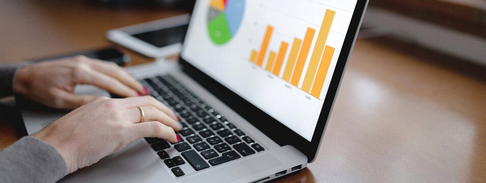 Horizontal banner close-up workplace with computer laptop and showing graphs on desktop with woman hands typing. Growth, analysis, tele working concept.
