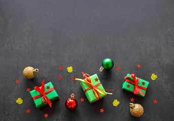 Gifts in green paper with red and yellow ribbons, gold, red and green christmas balls lie in a row. Confetti in the form of red stars are on the top. Black background, copy space. Big sale concept