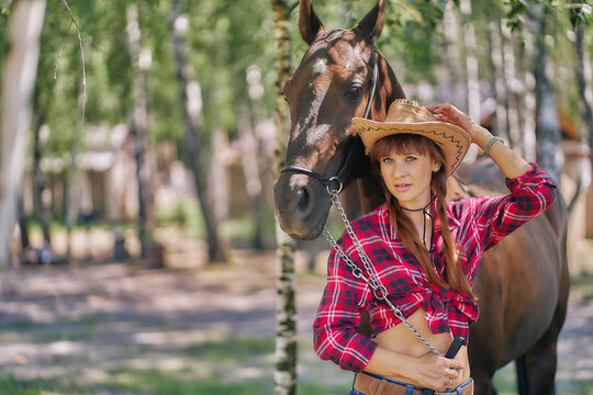 A woman in cowboy clothes holds a horse by the bridle and poses for a photo.
