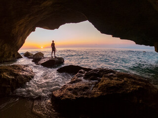 
Man from behind against the light inside a cave observes the sunset overlooking the sea, warm and...