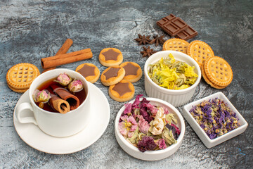 close up shot of a cup of herbal tea with cookies and cinnamons and dry flowers on grey background