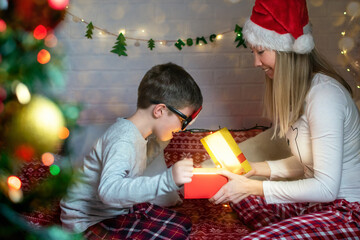 Obraz na płótnie Canvas Smiling mom in a Santa hat gives her son a Christmas present, the child is surprised, looks into the magic glowing box. Happy family opening gifts, New Year's Eve at home. Joy, emotions, festive mood