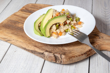 Quinoa porridge with vegetables and avocado in a white flat plate on a wooden board. Superfoods concept and top view.