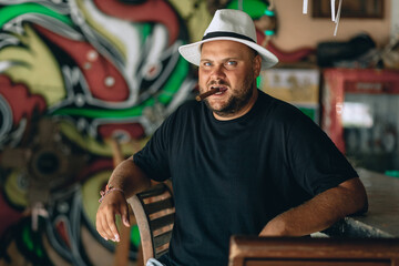 Portrait of a serious concentrated brutal man with a beard and a black and white panama hat on his head, holding a cigar in his teeth, looking at the camera.