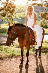  young blonde woman in a long white dress sits on a brown horse outdoors.