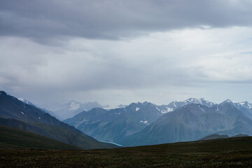 Fototapeta premium Gloomy view to great snowy mountains under dark gray cloudy sky. Dramatic alpine landscape with snow mountains in rainy weather. Atmospheric scenery with giant mountain ridge in overcast darkness.