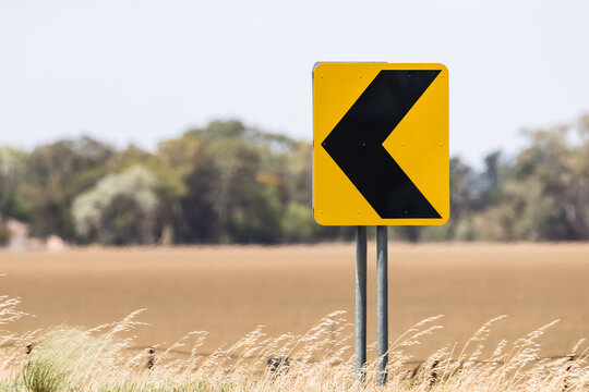 Black and yellow direction sign post on curve bend in road pointing from left to right with an empty field background in a rural remote area.  See also same image with sign pointing left to right.