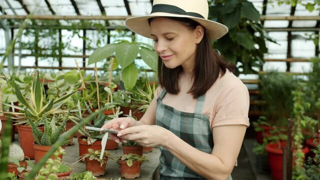 Young woman farmer is taking pictures of beautiful plants in greenhouse using smartphone camera advertising flowers in social media. People and technology concept.
