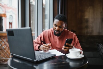 Profile portrait of young African online shop manager, talking on mobile phone to client, consulting him on delivery terms, sitting at cafe and using laptop computer for work, having coffee and cake