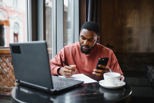 Profile portrait of young African online shop manager, talking on mobile phone to client, consulting him on delivery terms, sitting at cafe and using laptop computer for work, having coffee and cake