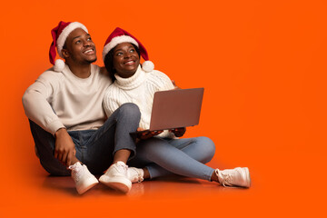 Dreamy african couple in santa hats sitting with laptop on orange background