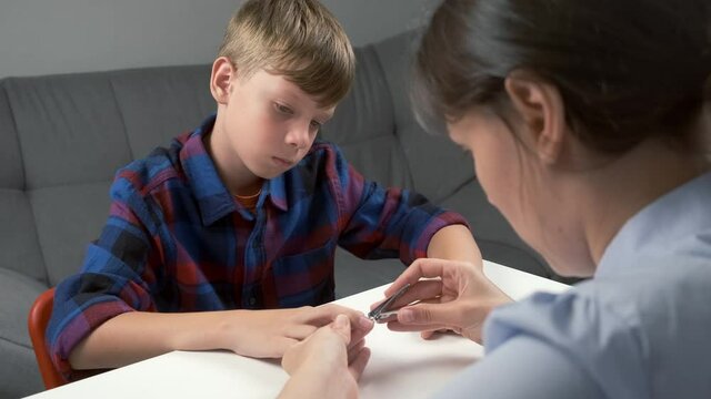 Mom is cutting nails of her son child boy using manicure nippers at home. Teenager is looking fo his hand. Mother manicurist master is doing manicure for him. Hygiene and care for hands of teenager.