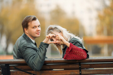 Fototapeta premium A young couple-brown-haired and blonde-sit on a bench and connect their fingers in the form of a heart