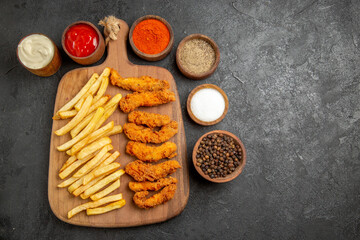Overhead view of fried chicken meal on cutting board served with differen spices on dark table