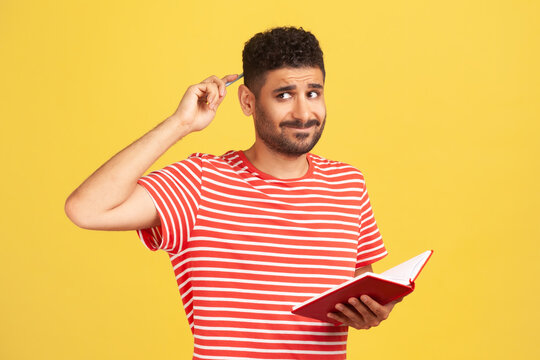 Upset Thoughtful Man In Striped T-shirt Rubbing Back Of Head With Pencil Holding Notebook In Hand, Writing Down Plans For Future, Making To Do List. Indoor Studio Shot Isolated On Yellow Background