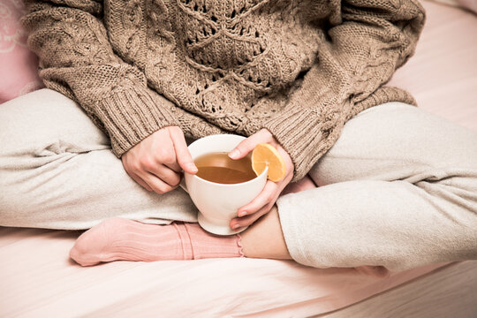 A girl young woman in a sweater and socks siting cross-legged sits in a sweater on the bed and holds a mug with hot tea and lemon. Lifestyle photo