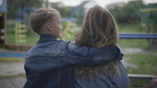 Positive Caucasian cute schoolboy hugging schoolgirl sitting on bench on playground. Back view of happy little couple resting outdoors on cloudy overcast spring or autumn day. First love.