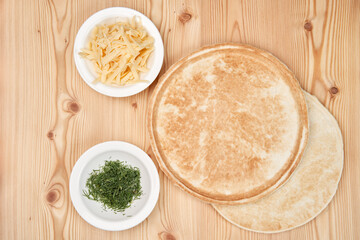 Two white plates with dill and cheese, and flat dough flat cakes for pizza on a light wooden background.