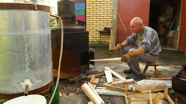 Man throws dry branches into the firebox of a homemade distillery making moonshine schnapps, alcoholic beverages.
