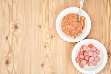 On a light wooden tabletop, white plates with chopped sausages and sauce. Copy space.