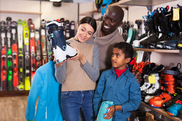 Fototapeta na wymiar Smiling young African man and European woman with preteen son choosing together ski boots for skiing in store of sports gear