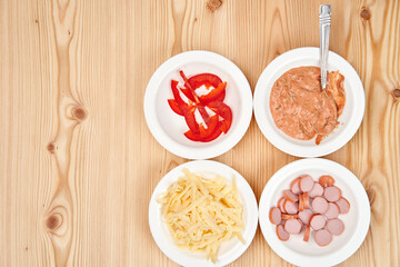 On a light wooden tabletop, white plates with chopped sausages, pepper, sauce and grated cheese. Copy space.