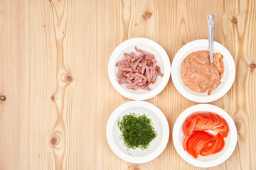 On a light wooden tabletop, white plates with chopped sausages, tomato, dill and sauce. Copy space.View from above.