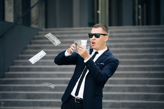 Successful business or winning the lottery. Money rain, falling dollars. Portrait shot of cheerful rich handsome businessman in glasses and suit throws money. Happy good looking man throwing dollars