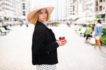 Elegant stylish blonde short hair girl in hat posing with coffee over street background