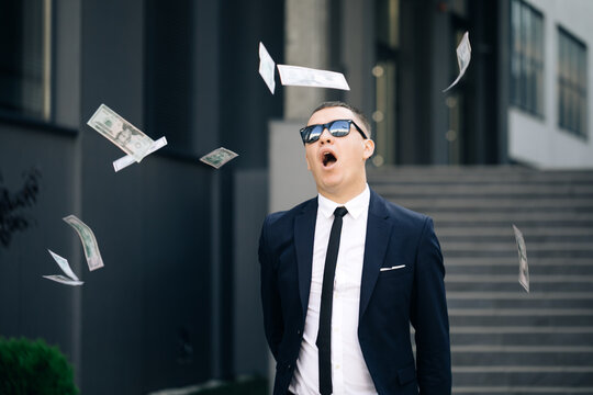 Happy good looking man throwing dollars. Money rain, falling dollars. Portrait shot of cheerful rich handsome businessman in glasses and suit throws money