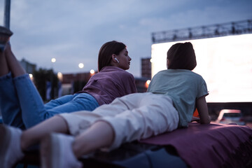 Two girls lying on the roof of a car, having fun while watching a movie in an open air cinema with a big white screen - obrazy, fototapety, plakaty