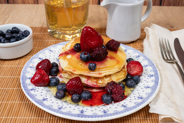 Stack of homemade pancakes prepared with blueberries, strawberries and maple syrup or honey dripping pancakes on an old plate on a wooden table. Concept healthy food at breakfast.