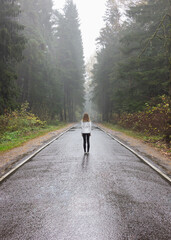 Girl Traveler in a white jacket stands on the road in a foggy forest.