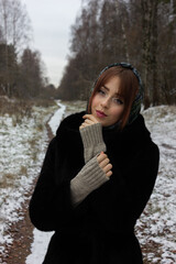 A Russian girl in a black fur coat and a green scarf in the winter looks at the camera.