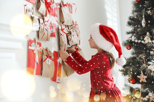 Cute little girl in Santa hat taking gift from Christmas advent calendar at home. Bokeh effect