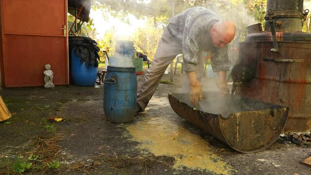 Man is transferring used fruit marc made of chopped apples, after distillation, using plastic bucket for filling small barrel to throw it as garbage. 