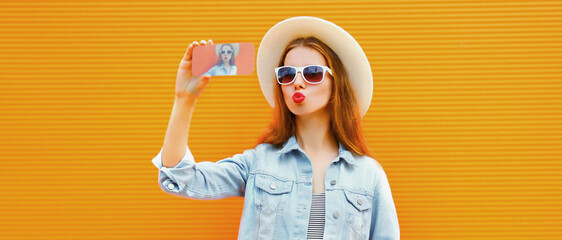 Portrait close up of young woman taking selfie picture by smartphone wearing a straw hat over an orange background