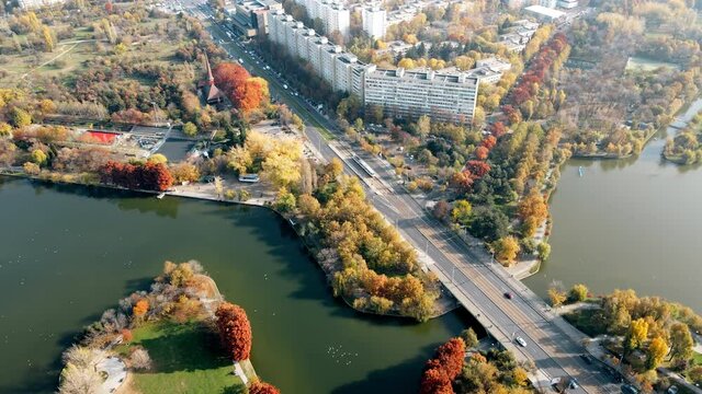 A road over the the lake with moving cars in the Titan park, multiple greenery and residential buildings. View from the drone, Bucharest, Romania