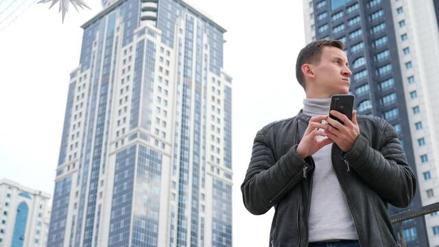 Concentrated handsome man finds best route using online maps on expensive smartphone walking against highrise buildings close low angle shot