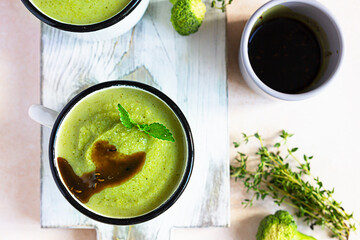 Two enamel mugs with healthy vegan broccoli soup with spicy oil and aromatic herbs over light background. Diet detox food concept. Top view.
