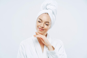 Pretty natural woman touches gently jawline, wears bath towel wrapped on head, dries hair after shower, cares about body, has clean skin, prepares for evening party, dressed in domestic robe