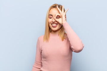 young blonde woman smiling happily with funny face, joking and looking through peephole, spying on...