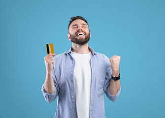 Excited young man with credit card gesturing YES on blue studio background