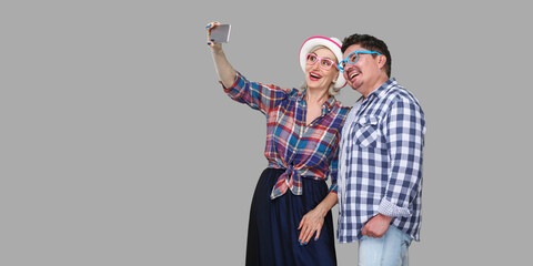Couple of friends, cute adult man and woman in casual checkered shirt standing together and making selfie foto for memory or video call, toothy smile. Indoor, isolated, studio shot, gray background