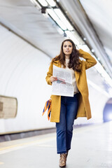 young woman in stylish autumn outfit looking at camera while holding newspaper on subway station